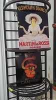 Two metal artwork pieces displayed on a metal rack, one for Vermouth Bianco Martini & Rossi and the other for Bitter Campari.