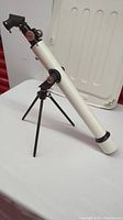 Full view of white vintage Tasco telescope mounted on a black tripod stand, placed on a white surface with a red background.