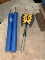 Photo showing two blue pool noodles attached to wooden sticks next to a yellow and grey Nerf axe.