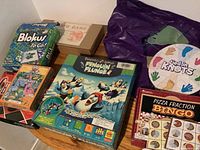 Photo showing collection of kids' games on a wooden surface including Penguin Plunge, Dinosaurs card game unopened, Blokus To Go, Pizza Fraction Bingo, Checkers, Earth Game boxed set, and Tied In Knots game bag.