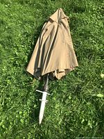 Closed sun umbrella with beige fabric and white ground screw base on green grass.