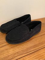 Side and angled view of black Kenneth Cole men's slippers showing moccasin-style toe and soft texture.