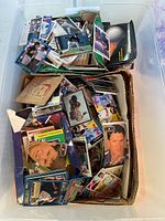 Top view of large plastic bin filled with assorted loose sports cards, visible are cardboard dividers and a mix of cards featuring athletes in baseball and basketball uniforms.