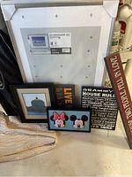 Photo showing multiple picture frames leaning against a wall including a large white IKEA frame, a framed scenic picture, and several small signs including a Mickey and Minnie Mouse sign and a Grammy's House Rules sign.