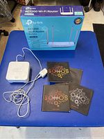 Photo showing TP-Link AC1200 WiFi router in sealed retail box on blue surface, with Sonos Bridge device, power cable, and manuals laid out.