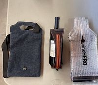 Photo showing a dark grey felt Mode two bottle wine bag with shoulder strap, a wine bottle in a brown stitched leather Graffigna holder, and six Wine Hero travel wine bags in bubble wrap