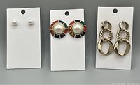 Three pairs of earrings displayed on white card stock: small pearl studs, large multicolor gold-tone earrings with central pearl, and twisted linked gold-tone drop earrings.