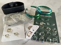 Photo showing various large silver-tone hoop earrings, two large green metallic hoop earrings, several pairs of small gold and silver tone earrings with some decorative stones, and one black rubber adjustable bracelet.