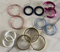 Top view of 8 pieces including rose gold hoop earrings, black hoops, pink sequin bracelets, blue hoop earrings, white hoops, and metal bangles on marble surface.