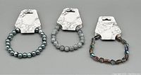 Photo showing three beaded bracelets on white marble display cards. One is metallic grey round beads, one is matte crackled light grey round beads, and one is faceted iridescent multi-colored beads.