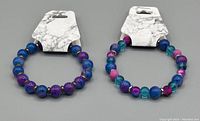Two stretch beaded bracelets on tag displays laid flat, colors including purple, blue, pink, and turquoise with gold accents on beads