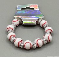 Image shows a handmade stretch bracelet consisting of white beads decorated with red baseball stitching and silver rhinestone spacer beads, packaged on a holographic card.