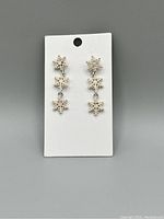Front view of a pair of silver-tone dangle earrings with three snowflake charms decorated with clear sparkly stones on a white display card.