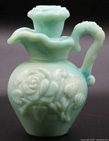 Front and side view of vintage pale green jade glass decorative pitcher with raised rose and leaf motif, showing scalloped rim and ornate handle.