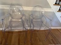 Front view of two clear acrylic chairs, showing oval backrest and armrests.