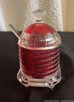 Front angle view showing beehive ribbed pattern of the glass mustard jar with clear lid, knob and spoon with red glass insert inside.