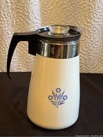 Full view showing white ceramic body, black handle, stainless steel top, and blue cornflower design on front.