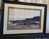 Framed print artwork showing a coastal scene with water, shoreline, trees, and a large reflective elongated object near the shore.