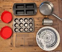 Top-down view of all items on wooden surface including Wilton silicone loaf pan, muffin pan, sifter, frying pan, serveware tray, cooling rack, and air fryer liners.