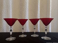 Front view of the set of four red top crystal cocktail glasses with clear stems lined up on a black surface.