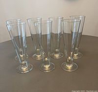 Seven clear pilsner beer glasses displayed on a gray surface, shown from an angled front view.