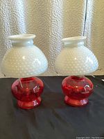 Pair of red and white hobnail milk glass hurricane oil lamps side by side on a black surface with textured beige background.