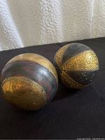 Two solid wood spherical decorative balls with brass inlay, one with a reddish wood base banded with engraved brass, the other with black wood base and ornate brass bands.