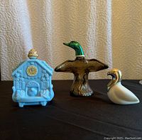 Three vintage Avon cologne bottles side by side: light blue cuckoo clock, brown flying mallard with green head, and white dove.