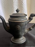 Front angled view of vintage pewter teapot with closed lid and emblem on body.