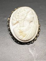 Close-up front view of a white carved cameo showing a woman's profile with floral details set in a twisted 10 kt yellow gold brooch frame.