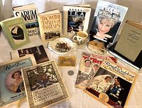 Group photo of memorabilia items including books, magazines, coin, cup, ashtray, and commemorative booklets on a white surface