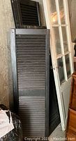 Photo showing a stack of black plastic shutters with realistic wood grain texture leaning against a stone/brick wall.