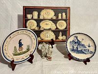 Main image showing entire lot: wooden display case with yellow-toned ceramic plates and jars, two standing plates on easels (Quimper Breton and Seaside Vista), and two porcelain figurines in front