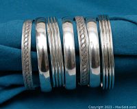 Seven silver bangles arranged horizontally on blue cloth, showing braided, smooth and ribbed designs.