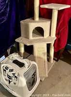 Front view of the beige cat tower with three carpeted platforms, one enclosed hideout, three sisal-wrapped scratching posts, and a carpeted ramp. Next to the tower is a white plastic litter box carrying case decorated with black cat illustrations and featuring a swinging door.