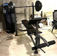 Full view of the assembled Marcy weight bench with barbell, showing leg extension rollers and preacher curl pad, against concrete basement walls.