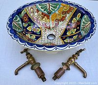 Oval ceramic sink showing colorful rustic blue, green, gold, floral/geometric pattern with royal blue scalloped border and central drain hole. Two brass faucet fixtures placed beneath.