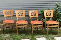 Four Stakmore wooden folding chairs in a row outdoors on grass. Each features pierced carved backrests and orange vinyl seats.