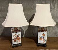 Pair of ceramic chinoiserie lamps with white fabric shades and black composite bases. Lamps feature hand-painted orange, green, and blue temple and floral motifs on white square bodies. Shades show some wear.