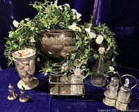 The overall lot with plated half-moon wall planter overflowing with green foliage and white flowers, large glass vase with shells, handblown bottle, two vintage silver plated caddies with cut and pressed glass cruets, and the sterling silver salt and pepper shakers at front left.