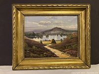 Front view of the framed oil painting depicting a landscape with a thatched hut near a lake and mountain background.