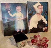 Photo of two classic portraits on canvas stretchers depicting people with birds and a small animal, a magenta Chalet art glass candy dish, and a floral papier-mâché planter.