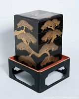 Front angle view of the four-tier lacquered jubako box on its black stand showing detailed gold bonsai tree design.