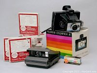 Photo showing vintage Polaroid Colorpack III camera in front of its original colorful striped rainbow box, accompanied by instruction manuals, Campbell Soup reel to reel tapes boxes, and a pack of flash cubes.