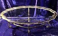 Full overview of the oval coffee table showing brass scalloped rim, clear thick inset glass top, and curved brass legs with scrolled feet on blue fabric background.