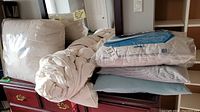 Two duvets stacked alongside several white pillows on a wooden chest with two unopened pillowcase packages on top.