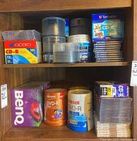 Shelf holding multiple brands of blank DVD and CD discs in boxes, spindles, and cases, showing brand names Memorex, Maxell, Cicero, Verbatim, and BenQ.