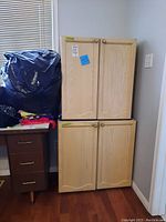 Photo of two stacked upper kitchen cabinet units next to a window and side table with clothing and bags on top