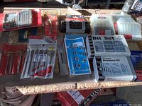 Overview image of various drill bit sets and accessory kits arranged on a surface including red case set, multiple packaged sets, and accessory items