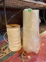 Photo showing an incomplete wooden spool of white rope and a partly unrolled transparent plastic bag roll placed on a carpet near a metal structure, illustrating lot contents.